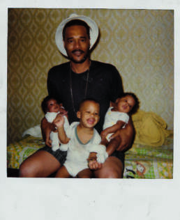 Black man holding three babies - one in each arm and one is on his lap.